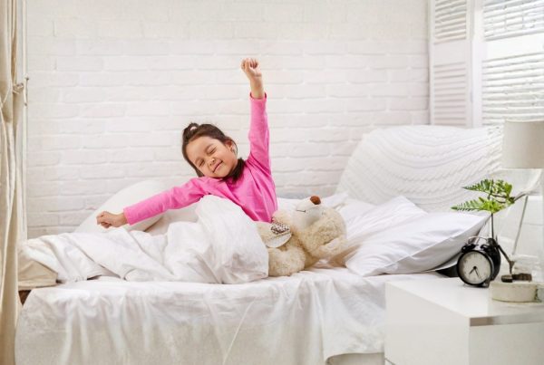 Child stretching and smiling while sitting up in bed in a bright bedroom, with bedding and a bedside table visible, representing a typical morning routine in a home setting.