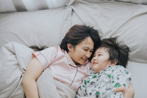 Parent and young child lying together in bed, smiling and cuddling, illustrating a supportive family environment around childhood sleep and nighttime routines.