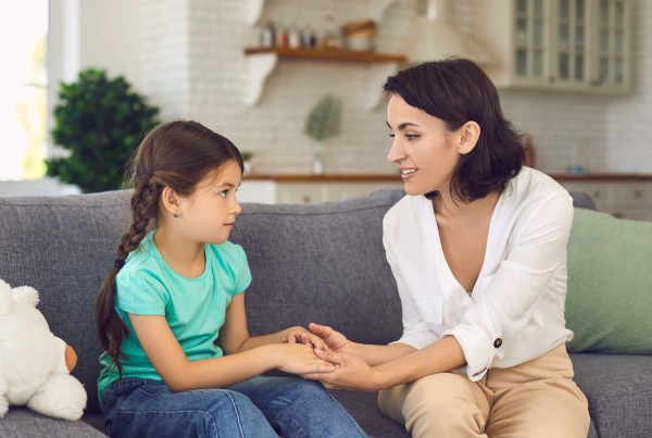 Parent sitting on a sofa holding a young girl’s hands while talking with her in a bright, comfortable living room.