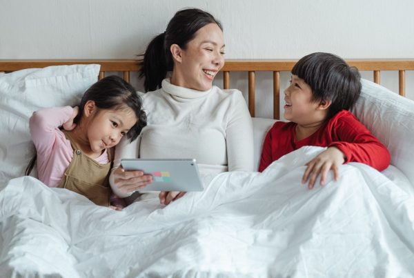 A mother sits in bed with her two young children, sharing a lighthearted moment while looking at a tablet together and discussing Helping siblings understand and support each other