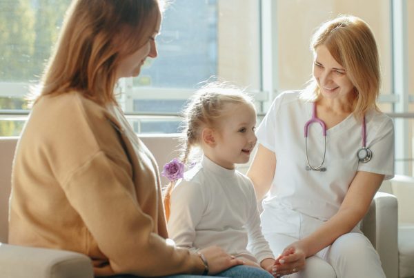 A smiling female doctor talks gently with a young girl and her mother during a supportive medical consultation.